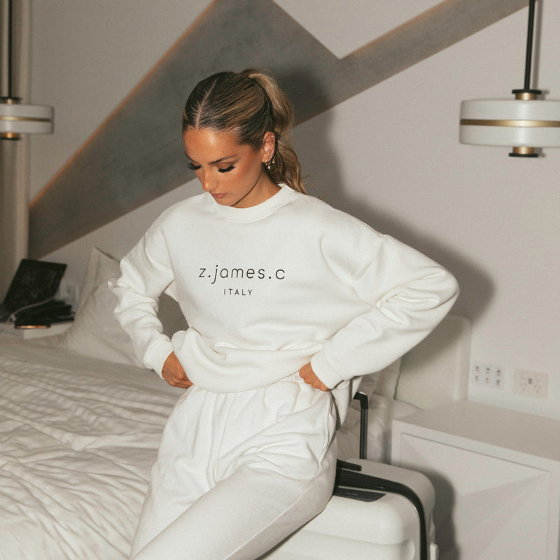 A female model wearing a white sweatshirt by z.james.c. She is looking down towards the ground with her hands on her hips.